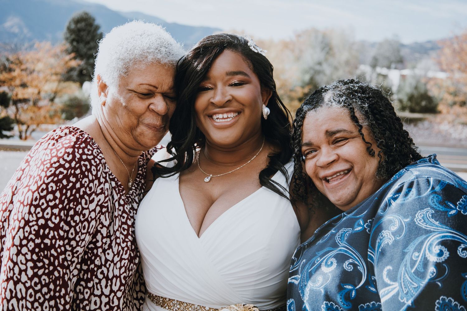 Bride in white wedding dress smiling with two women outdoors with mountains in background.
