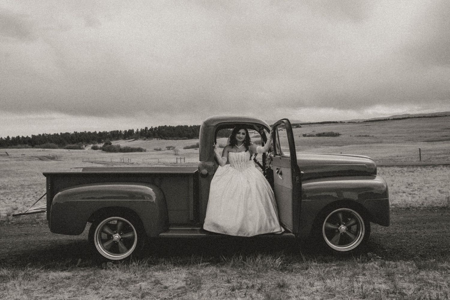 Bride in white gown posing beside a vintage pickup truck in a black and white photo.