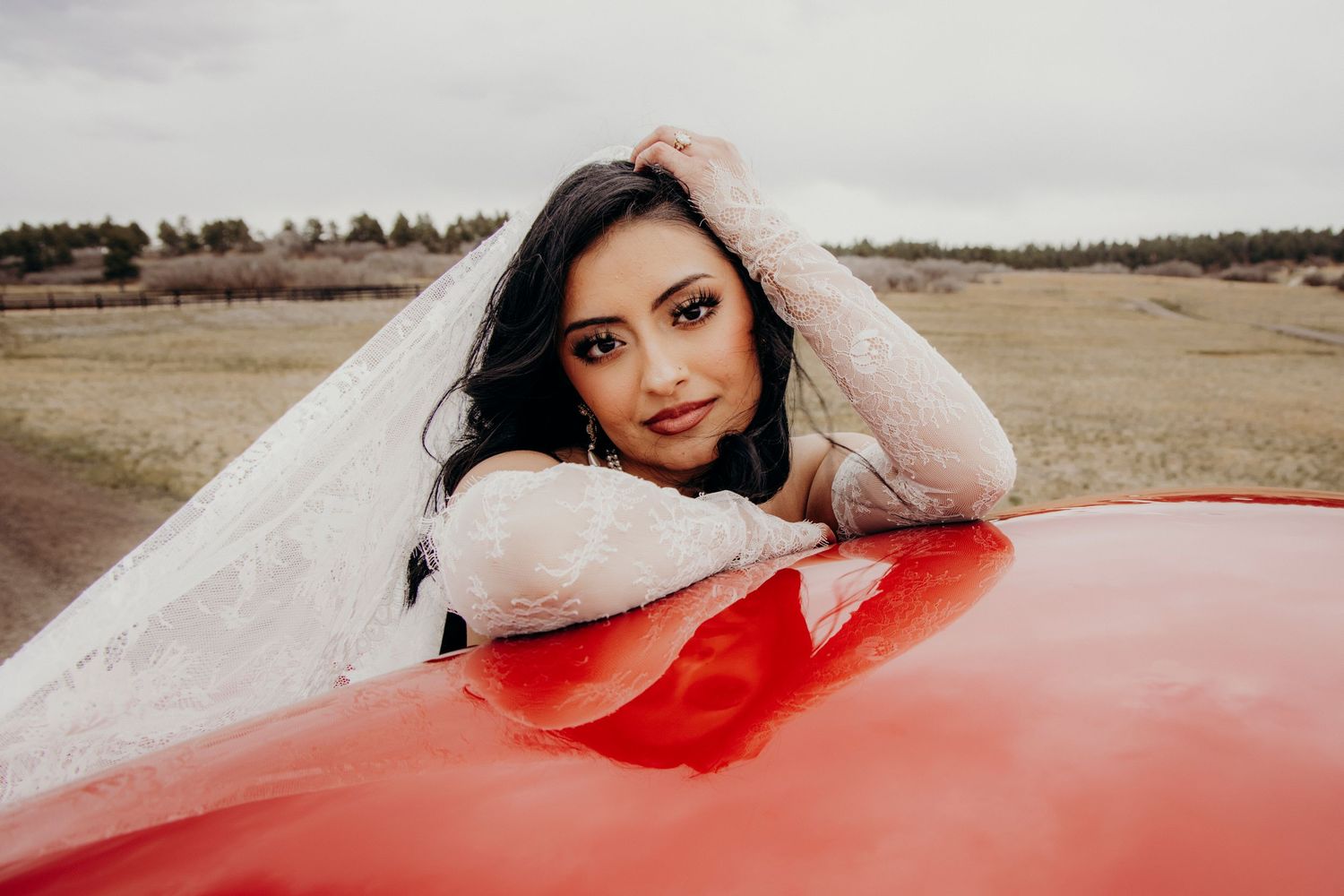 Beautiful bride in white lace dress leaning on a red vintage car in an open field.