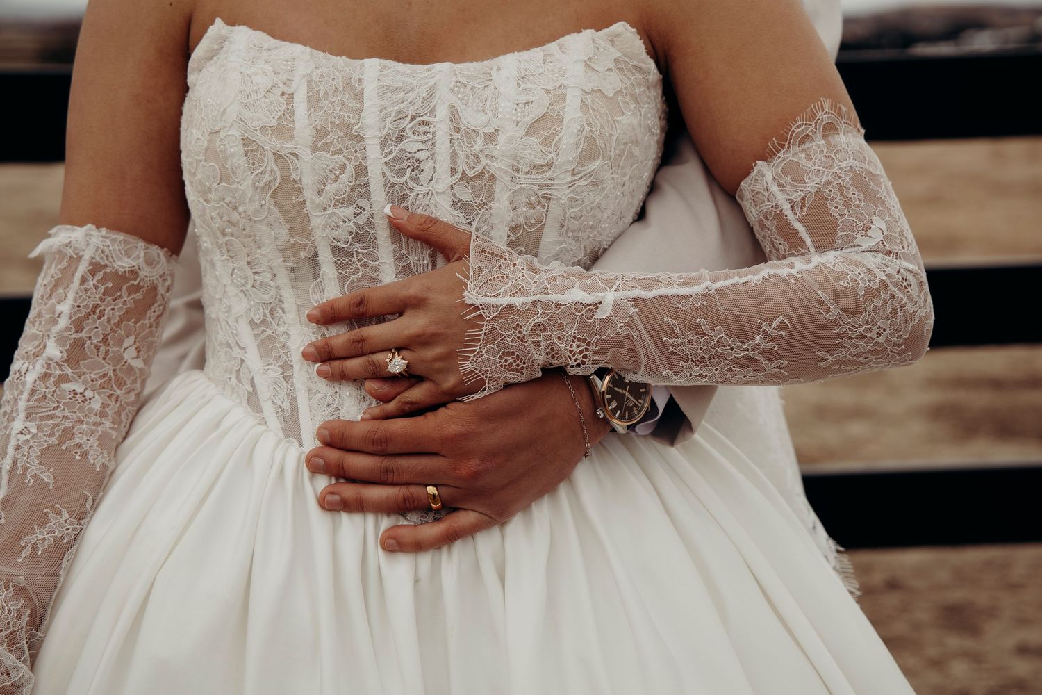 Groom embracing bride from behind showing wedding rings and lace bridal gloves.