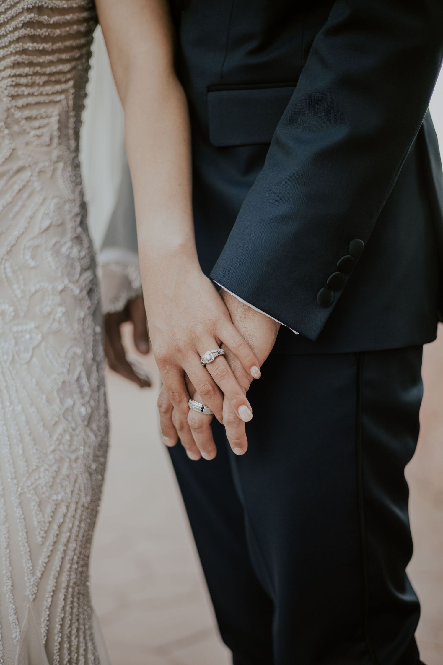 Bride and groom holding hands showing diamond wedding rings, bride in lace gown, groom in black suit.