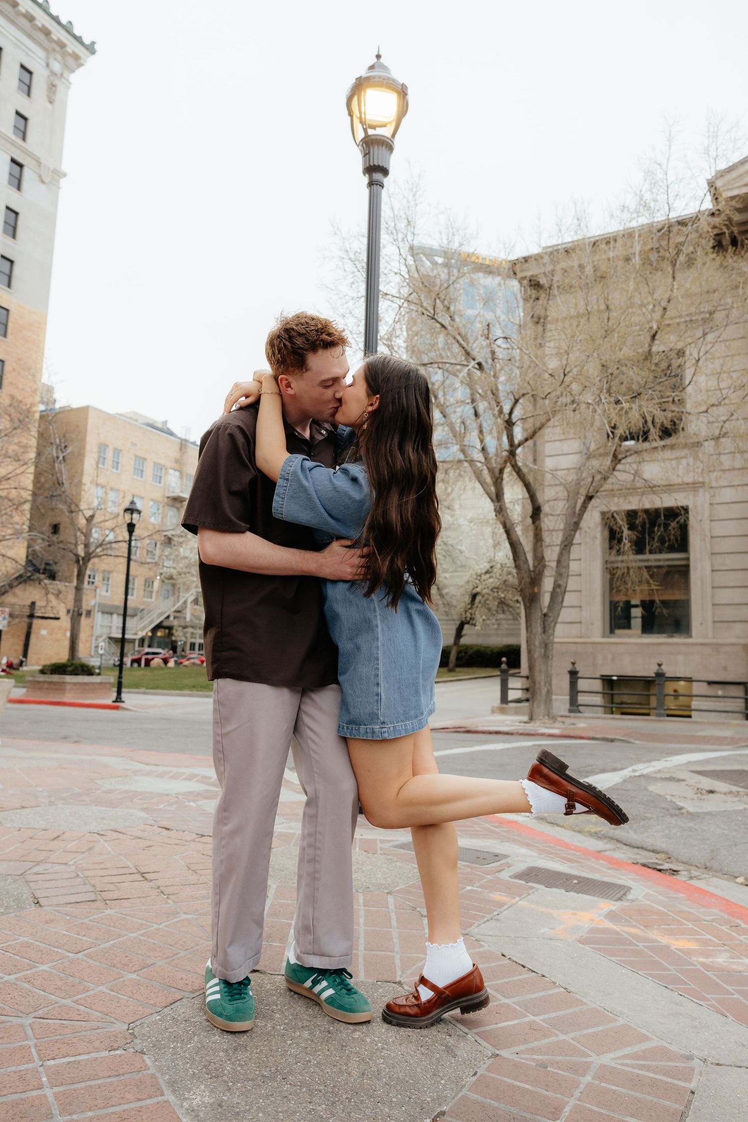 Young couple sharing a passionate kiss on urban brick plaza beside a glowing street lamp in spring.