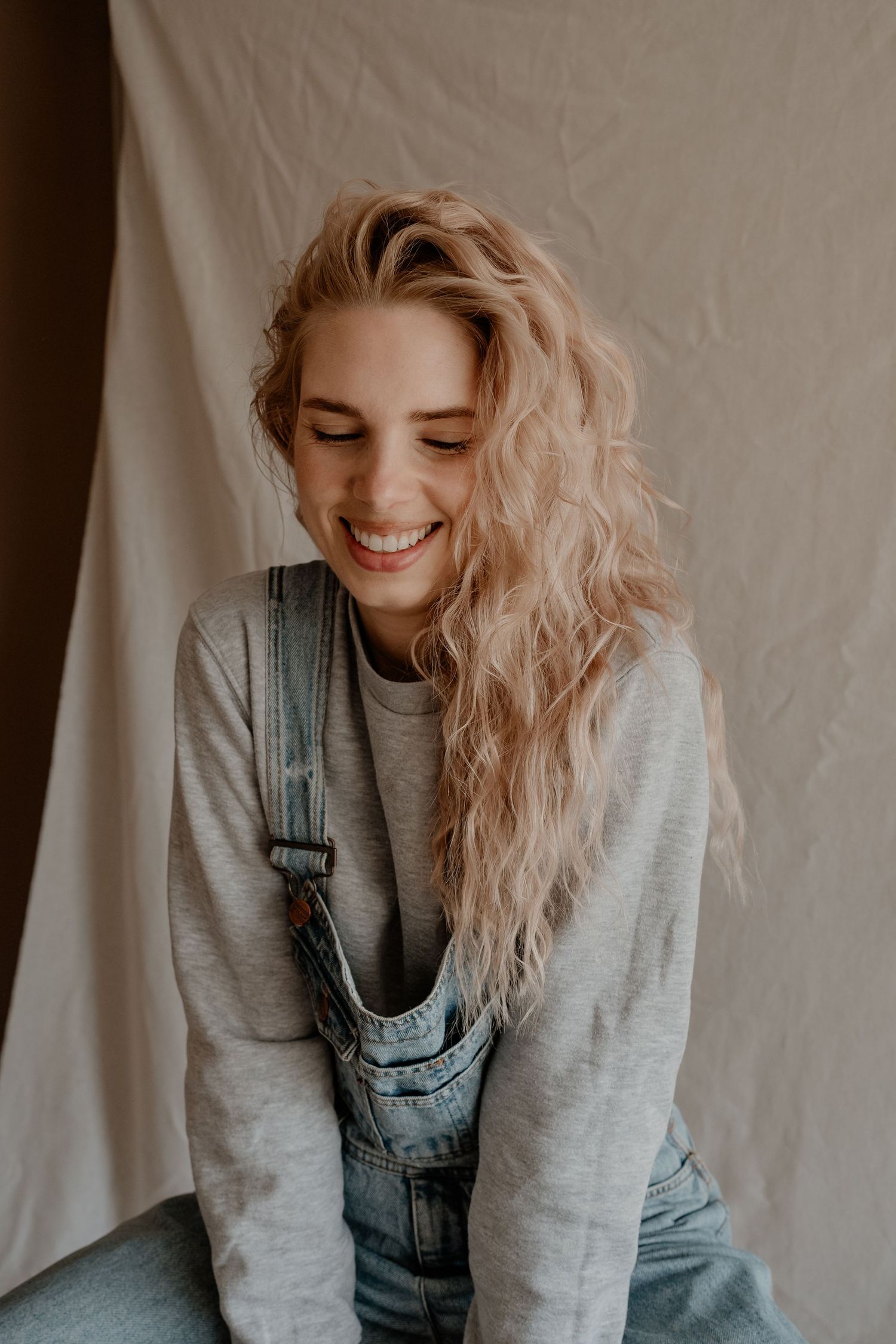 Persoon met golvend blond haar, gekleed in een grijze trui en denim tuinbroek, glimlacht met gesloten ogen tegen een neutrale achtergrond.