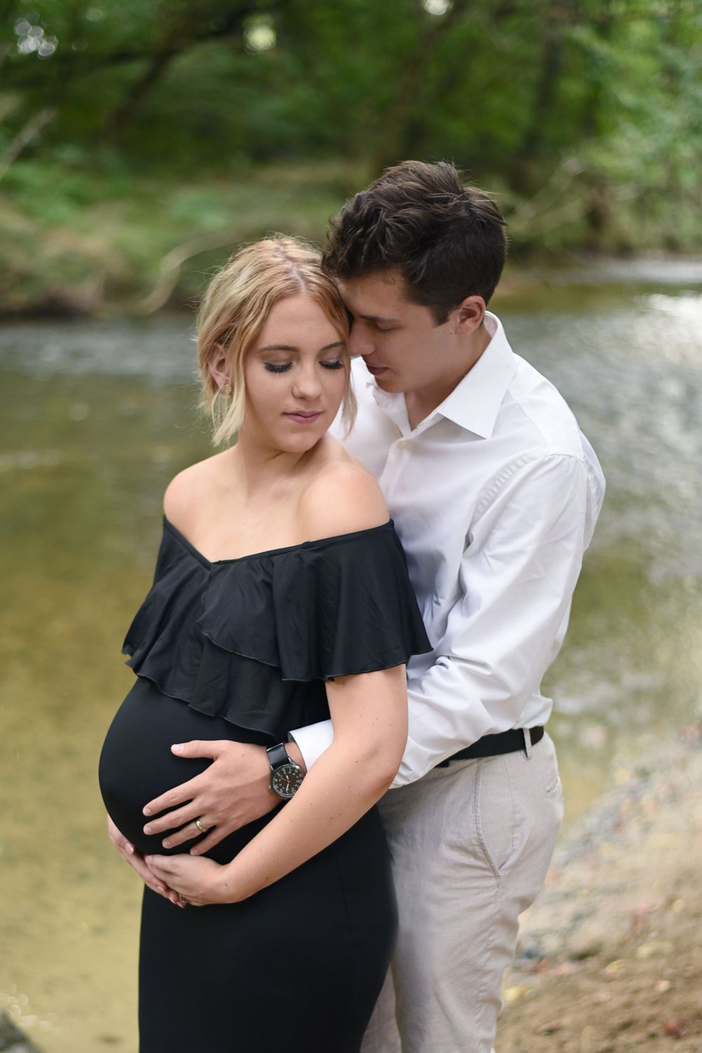 Maternity photos by water showing couple in black dress and white button-down shirt.