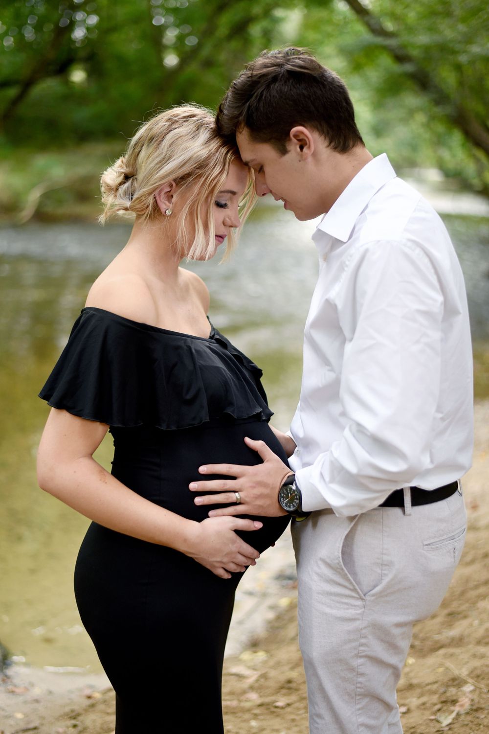 Intimate maternity photoshoot by a lake with couple in black dress and white shirt.