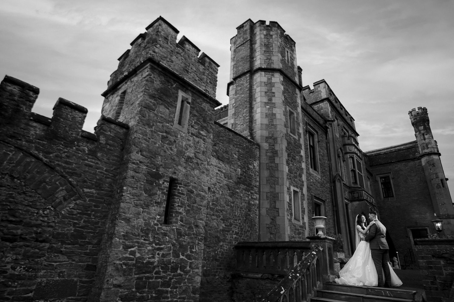 A bride in a white fitted dress and veil walks along a garden path while her groom follows behind in a black suit.