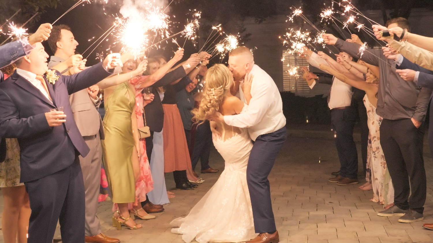 Newlyweds kiss during a festive night sparkler exit with cheering guests.