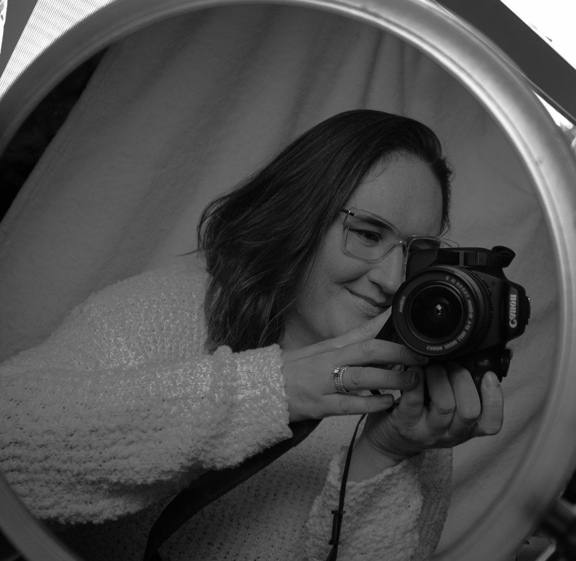 Black and white photo of photographer Hannah holding a camera while taking a self-portrait in a mirror.