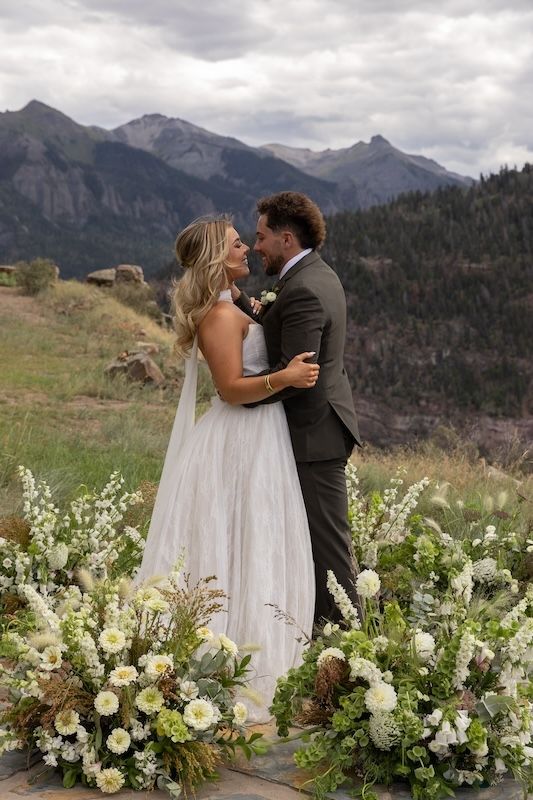 Bride and groom embrace at mountain wedding altar surrounded by white and green floral arrangements with scenic peaks backdrop.