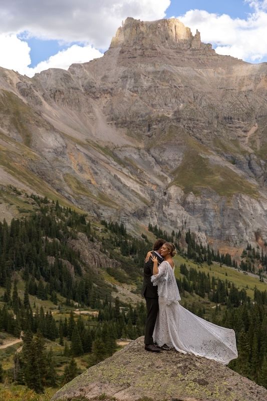 Bride and groom embrace on a rocky cliff with stunning mountain peaks and evergreen forest backdrop.