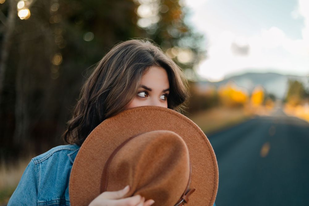 Someone peeks out from behind a brown fedora hat while wearing denim on a rural road at sunset.