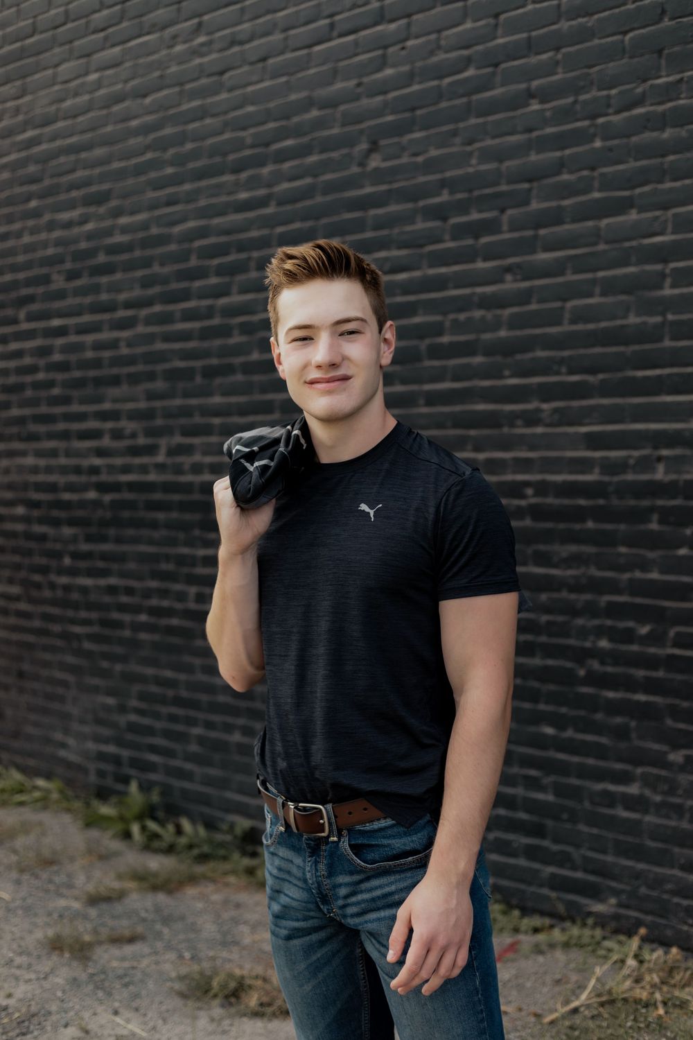 A photographer holding a camera poses against a black brick wall while wearing a dark t-shirt and jeans.