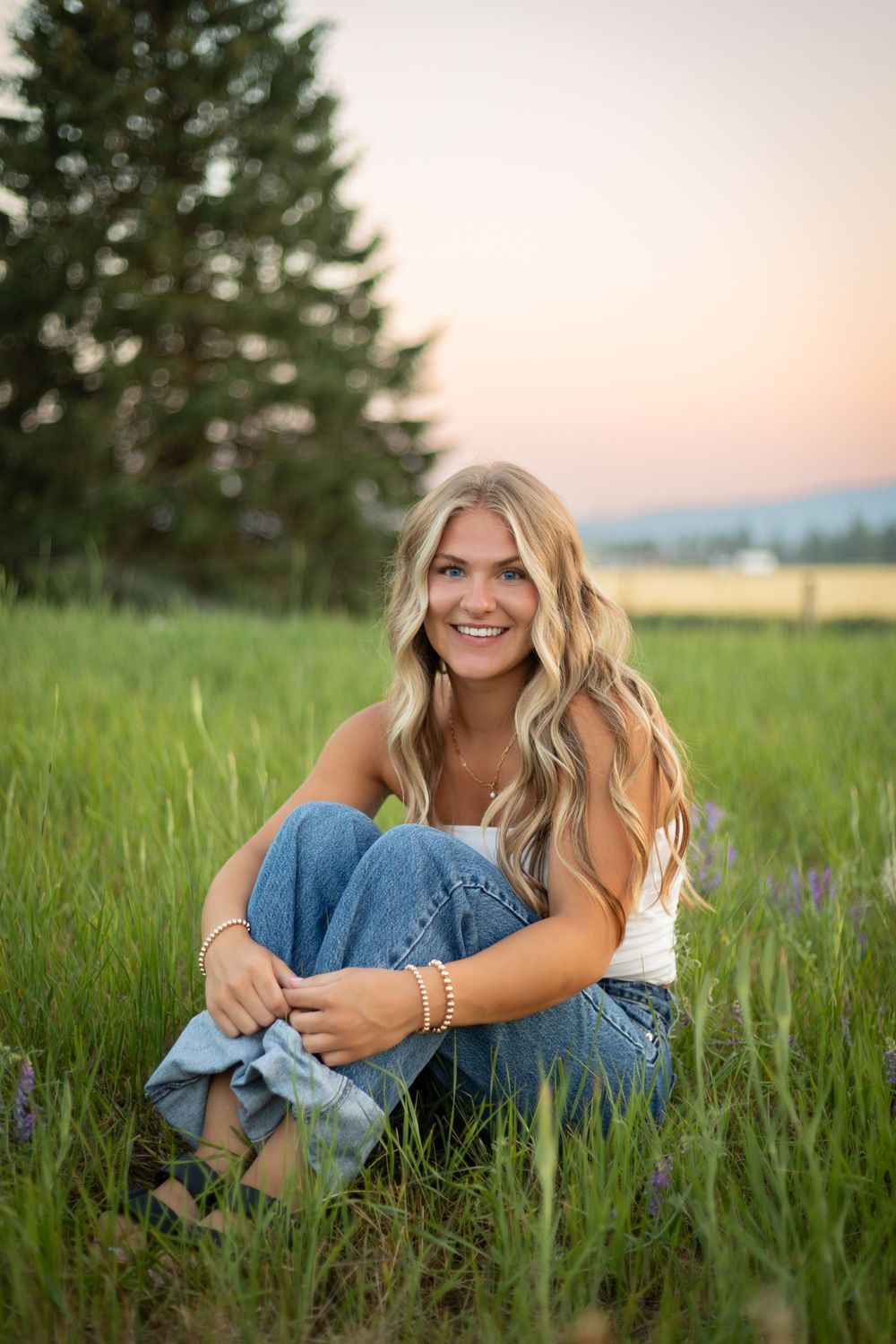A person sits in a lush green field at sunset wearing jeans and a white top with mountains visible in the background.