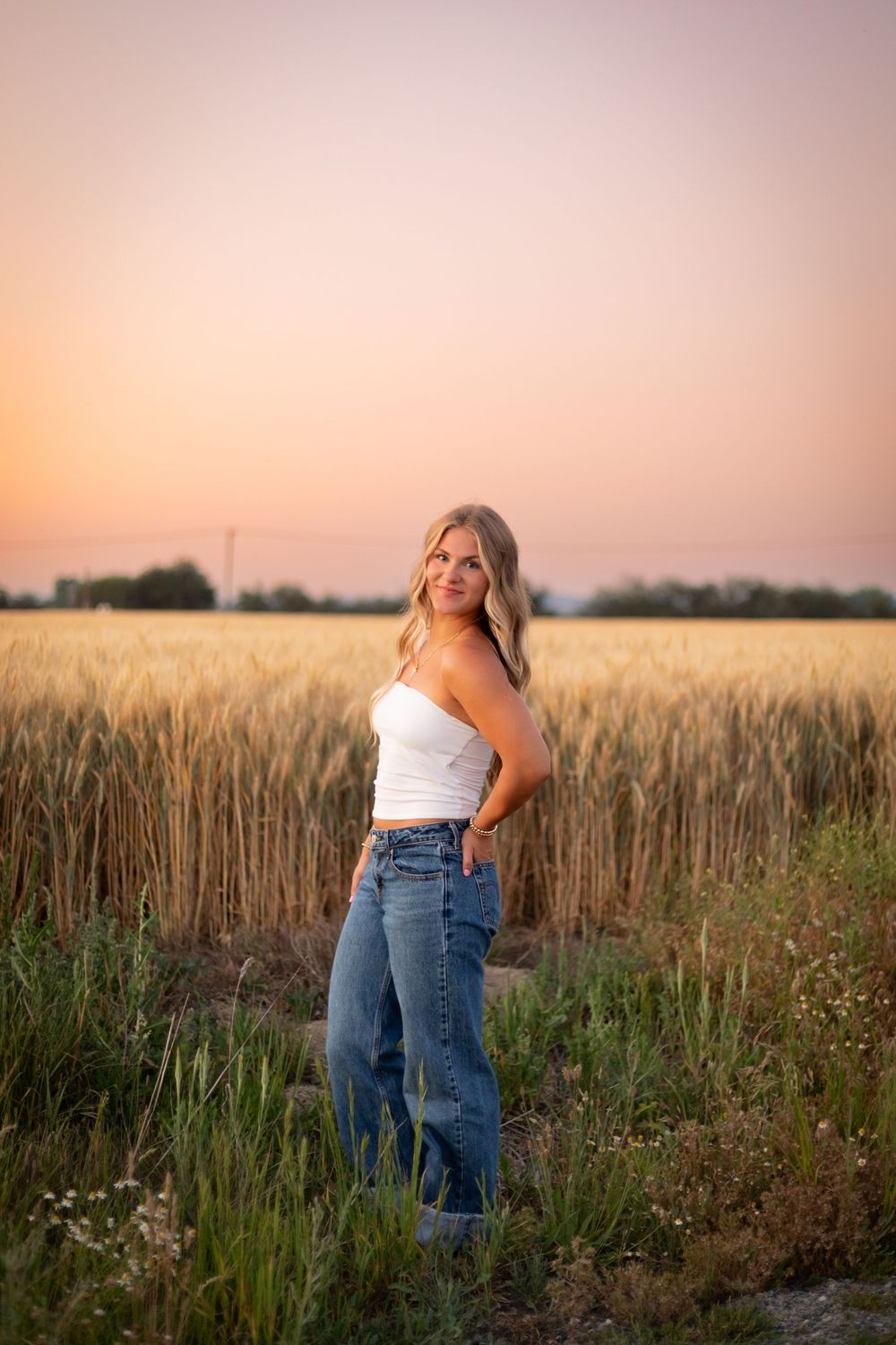 A casual summer outfit with white tank top and jeans in a golden wheat field at sunset.