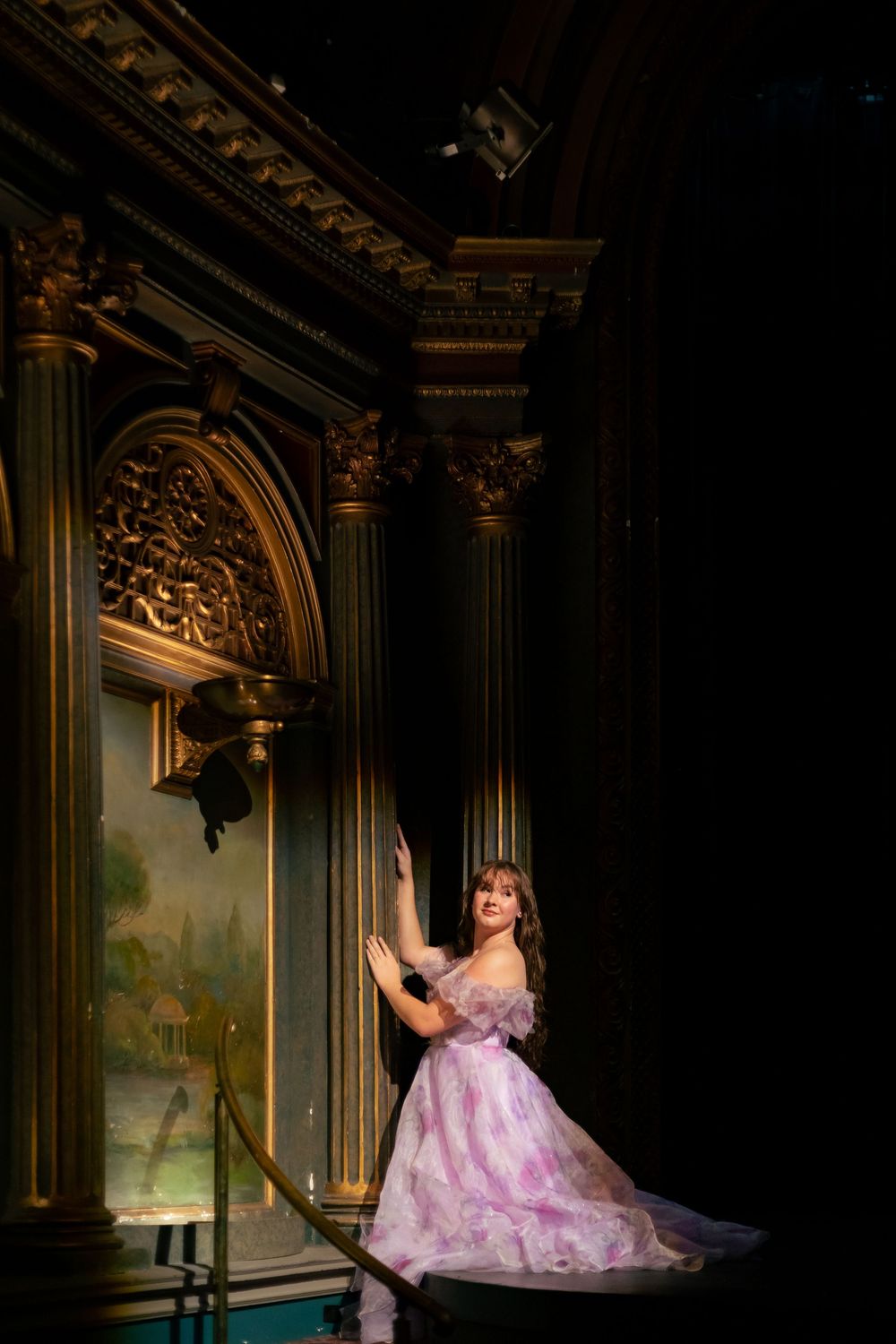 A dancer in a flowing pink gown poses elegantly against ornate architectural details in a dramatic theatrical setting.