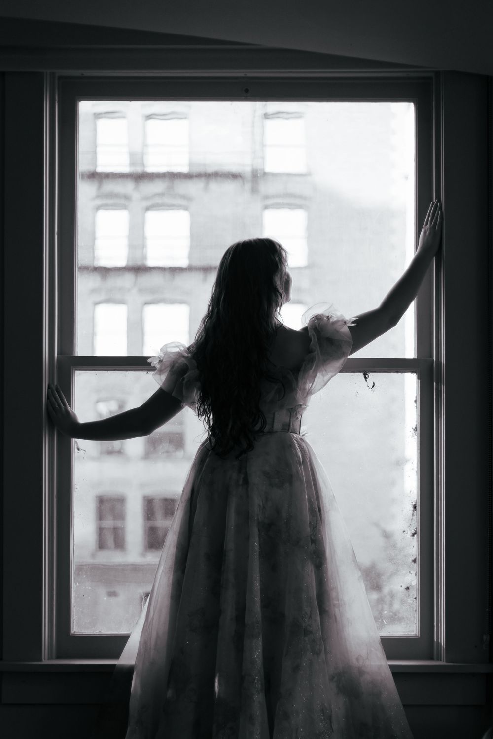 Silhouette of a figure in a flowing dress stands at a window with arms outstretched against city buildings.