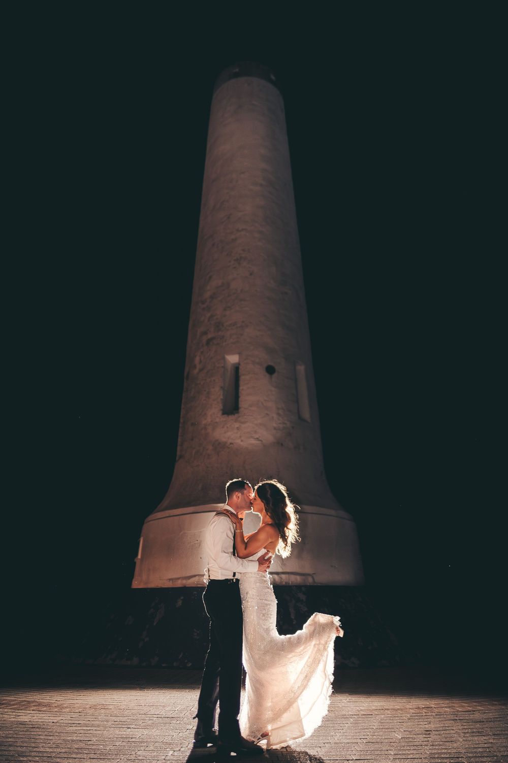 Bride and groom share a romantic kiss at night in front of an illuminated stone tower.
