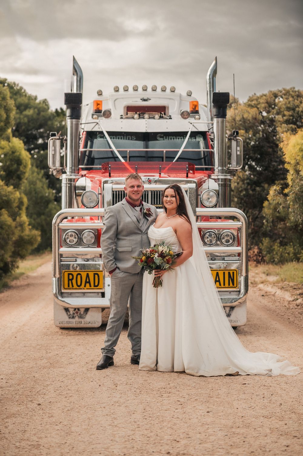 Bride and groom pose in front of a large semi-truck on a dirt road on their wedding day.