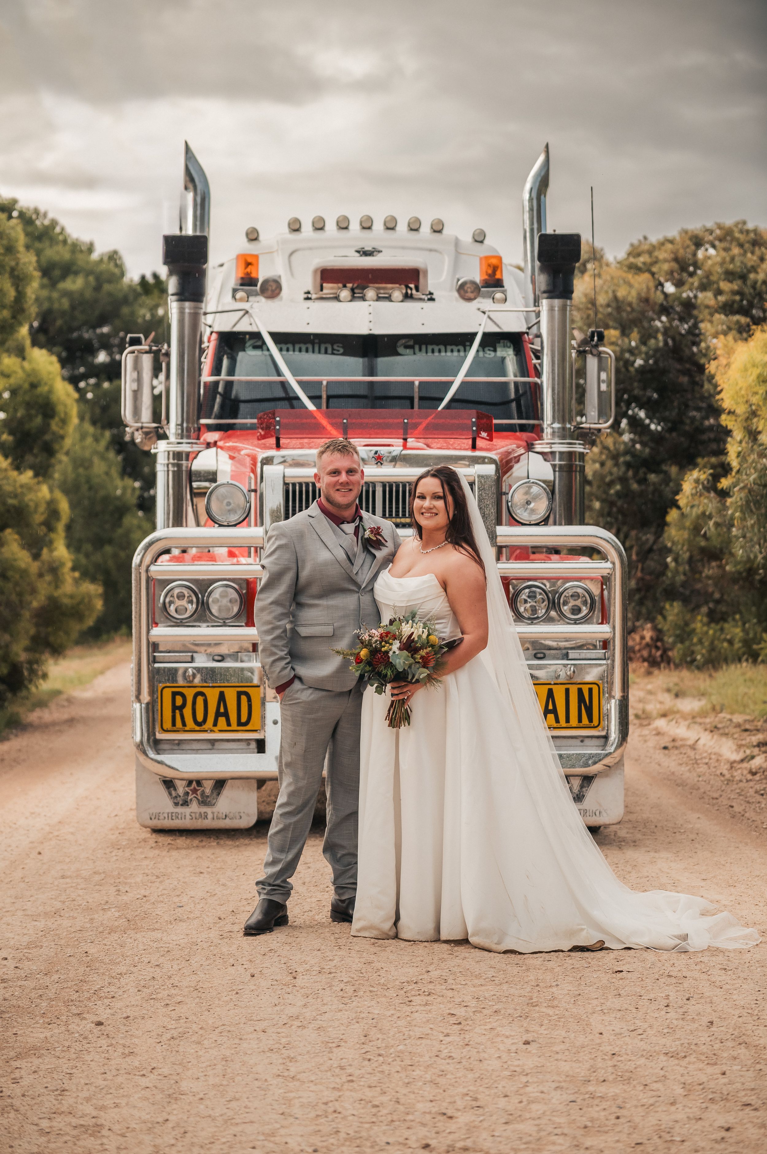 Bride and groom pose in front of a large semi-truck on a dirt road on their wedding day.