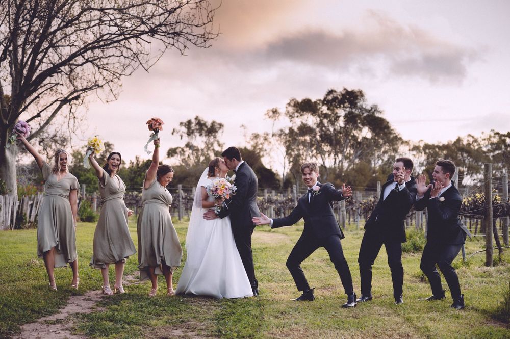 Wedding party celebrating outdoors at vineyard, bride and groom kissing while bridesmaids and groomsmen cheer around them.