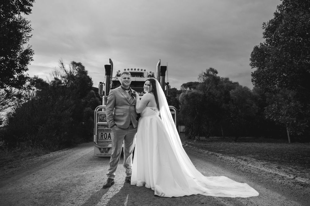 Black and white photo of a bride and groom posing in front of a vintage truck on a rural road.