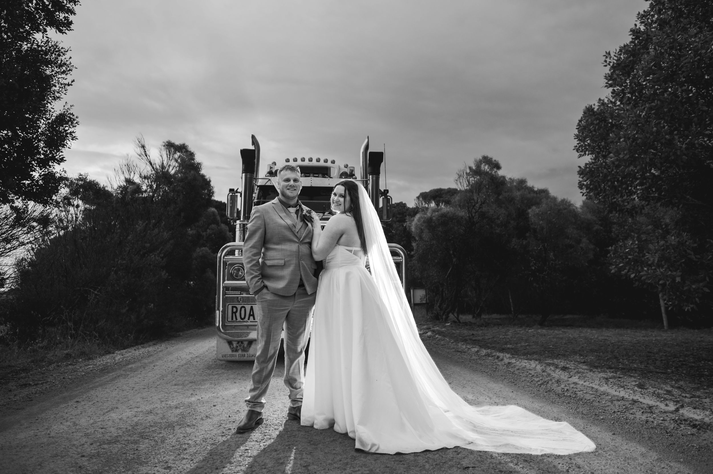 Black and white photo of a bride and groom posing in front of a vintage truck on a rural road.