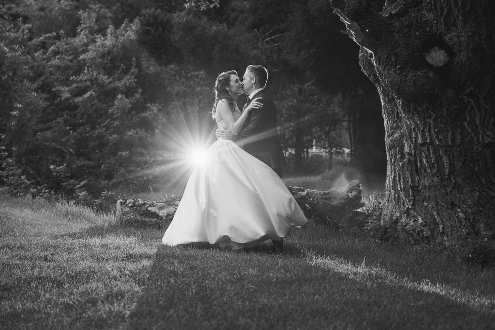 Black and white wedding photo of bride and groom kissing outdoors with sunburst light effect near a tree.