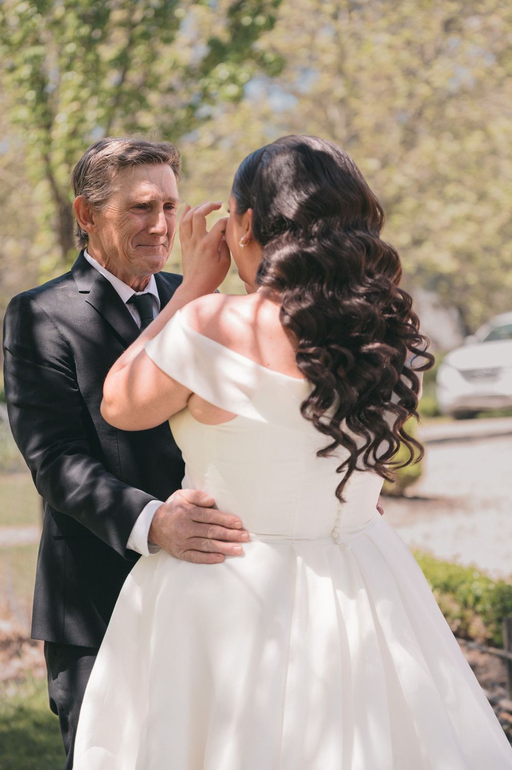 Emotional father wiping tears seeing bride daughter in white off-shoulder wedding gown outdoors.