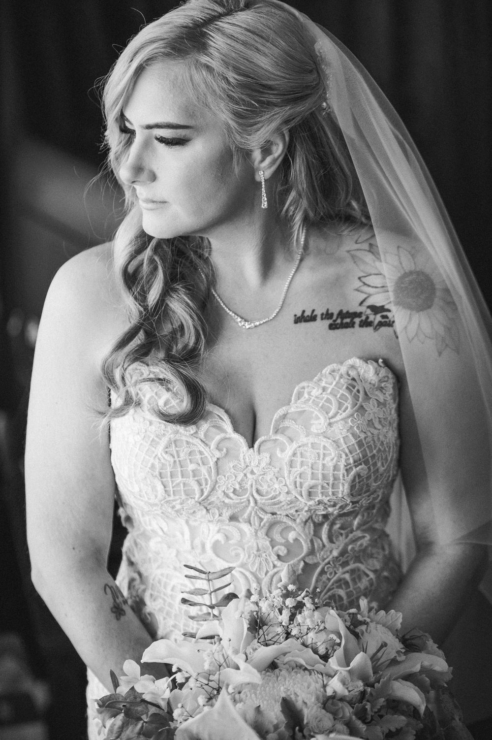 Black and white bridal portrait of a tattooed bride in lace strapless gown with veil holding bouquet.