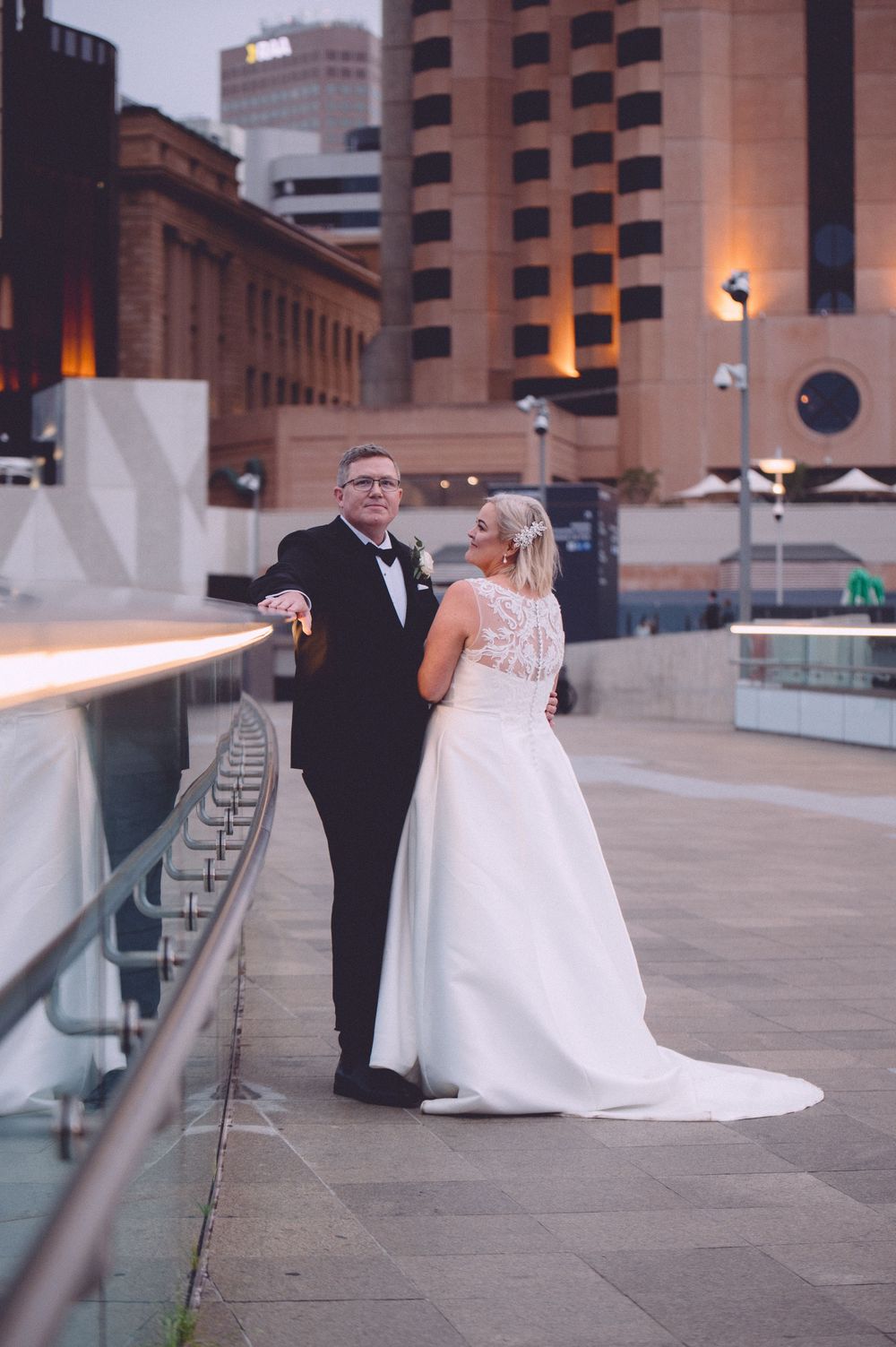 Bride and groom pose on urban rooftop terrace at dusk, bride in elegant white lace gown, groom in classic black tuxedo.