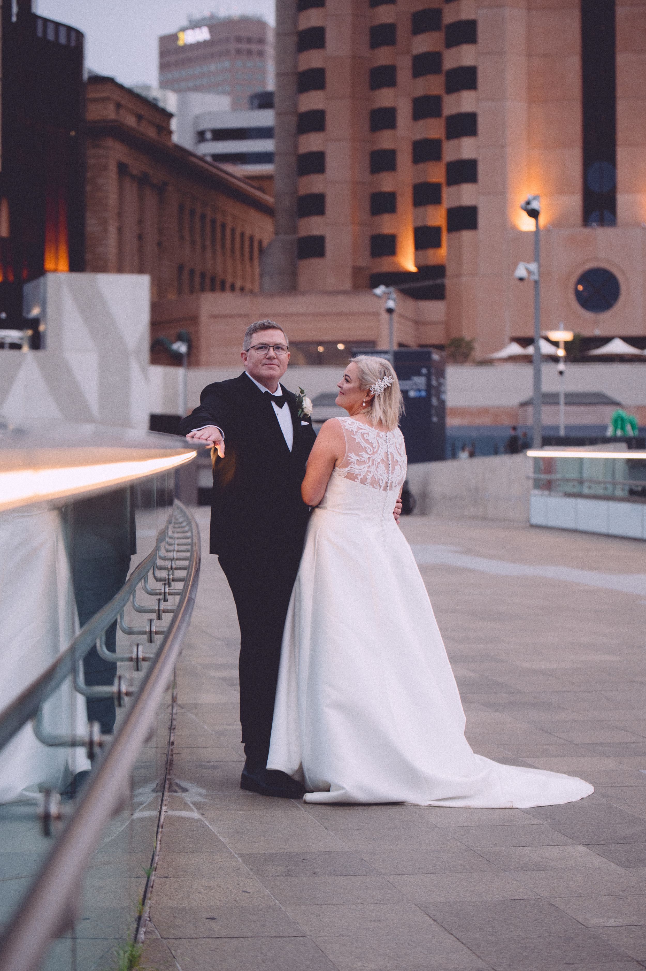 Bride and groom pose on urban rooftop terrace at dusk, bride in elegant white lace gown, groom in classic black tuxedo.