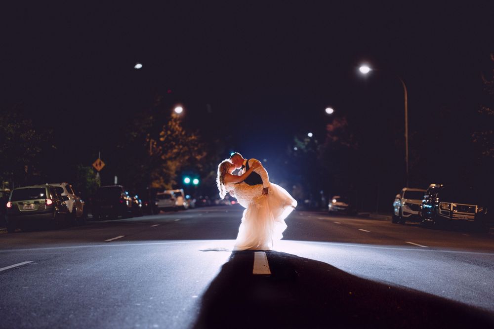 Couple in wedding attire sharing a romantic dip kiss on a city street at night, illuminated by street lights.