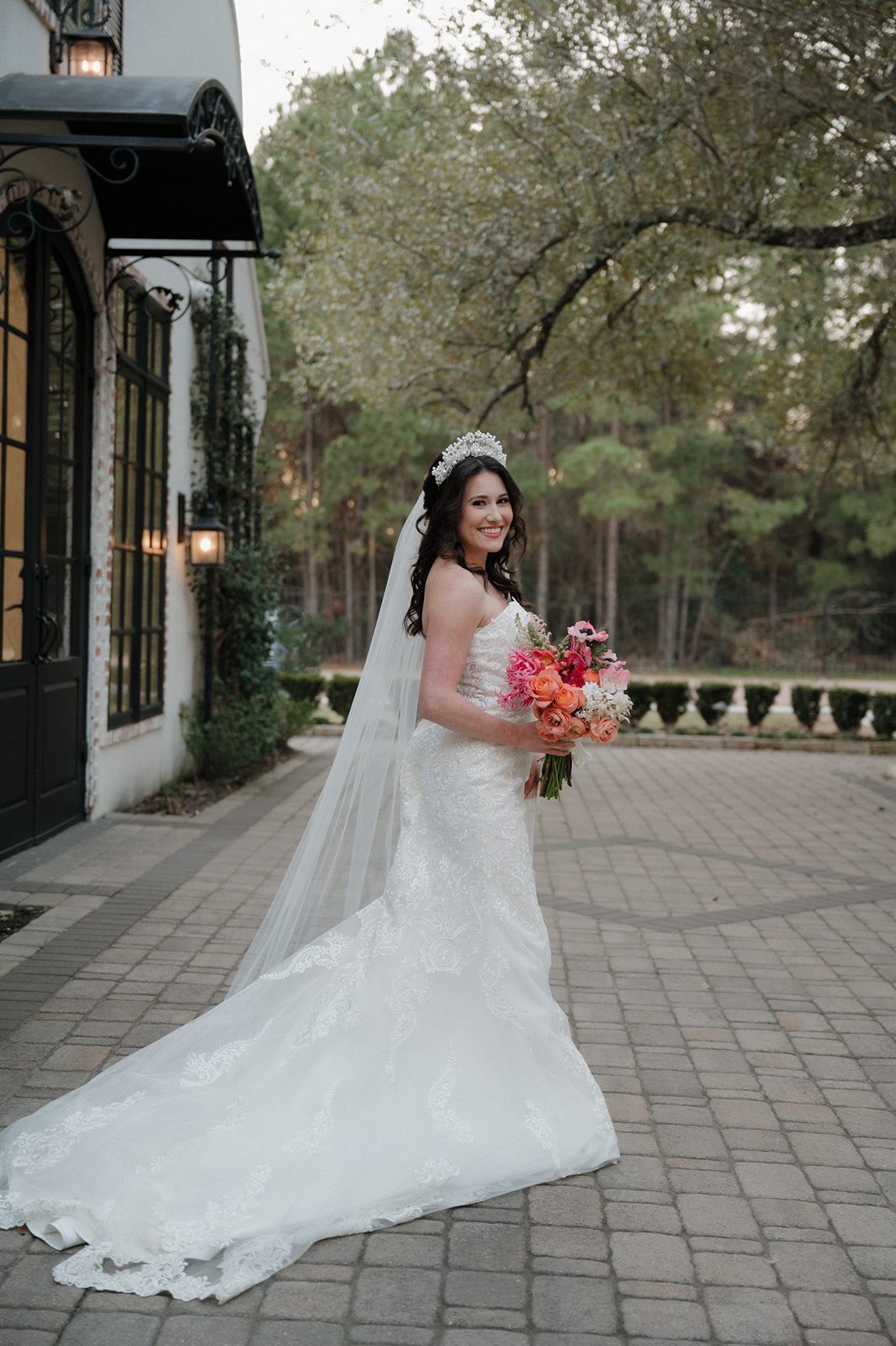 Beautiful bride in white lace gown with long train holds colorful bouquet by elegant building on brick patio.
