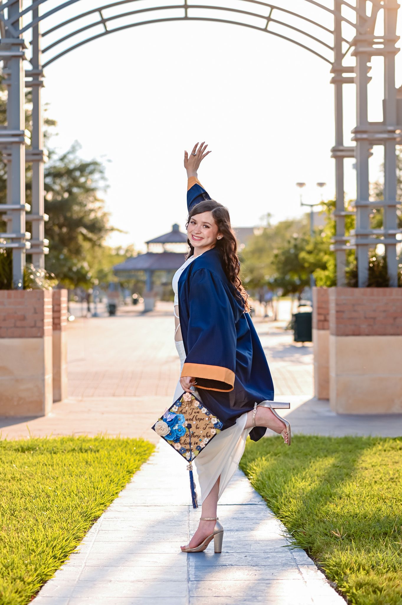 A graduate in a navy blue gown poses playfully under an archway.
