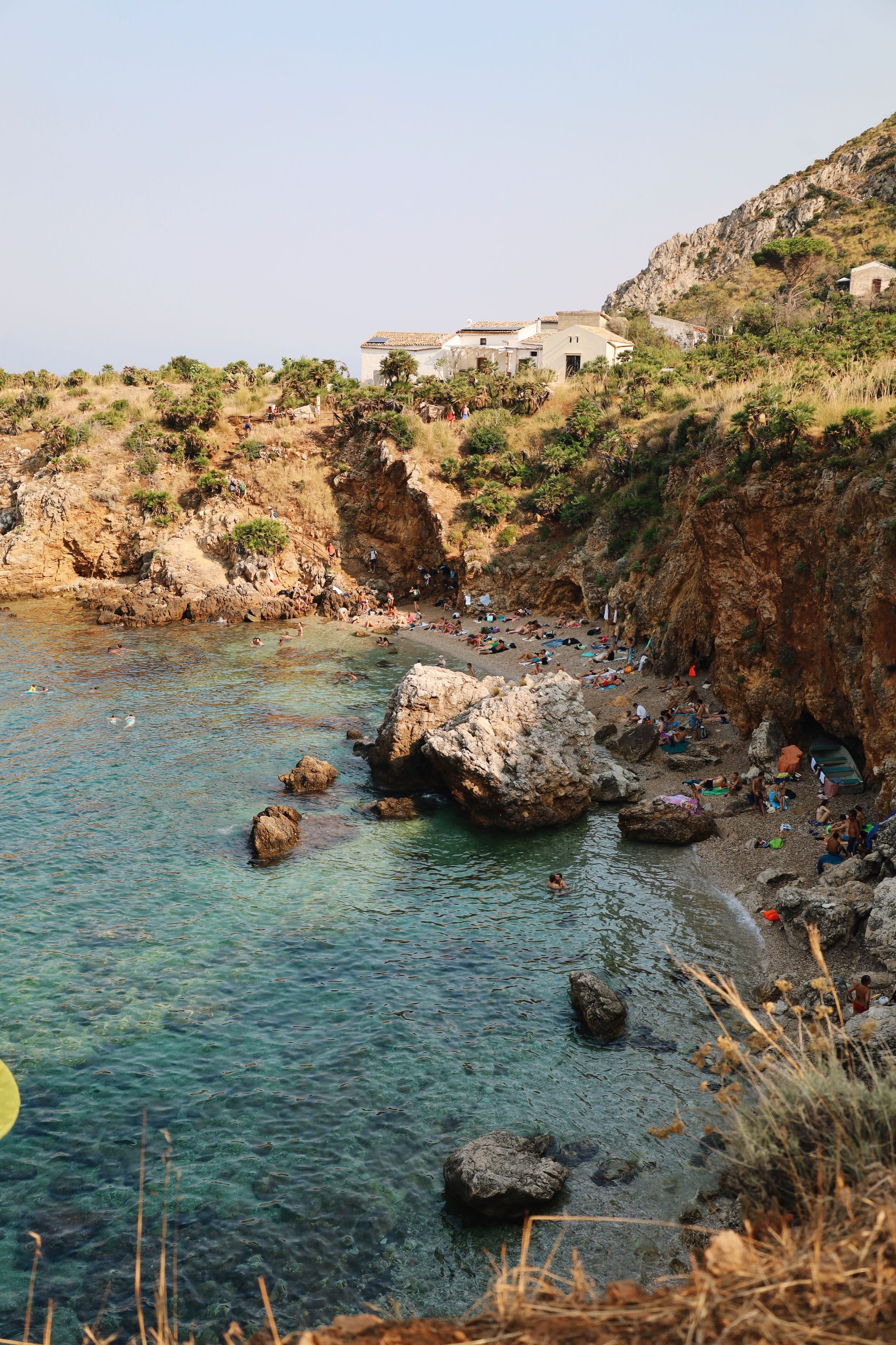 Crystal clear turquoise waters meet rocky cliffs at a Mediterranean beach cove filled with beachgoers.