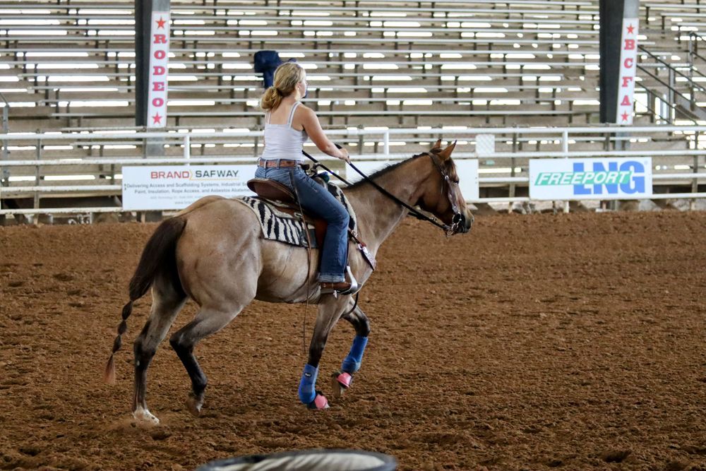 Riders on horseback compete in a rodeo event inside a dirt arena with bleachers.
