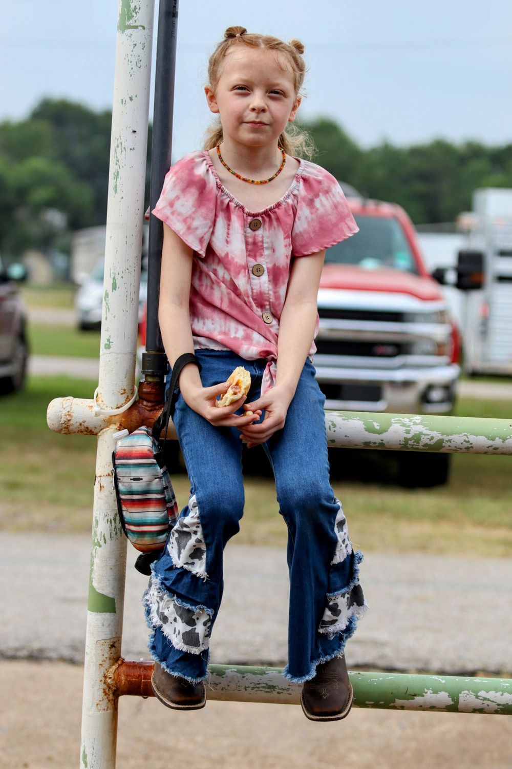 Young person in pink shirt and jeans sits on metal fence at outdoor event.