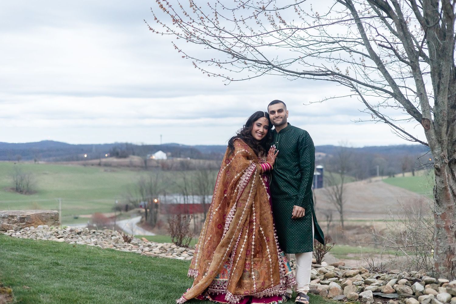 Couple in traditional wedding attire posing outdoors with rolling hills in background.