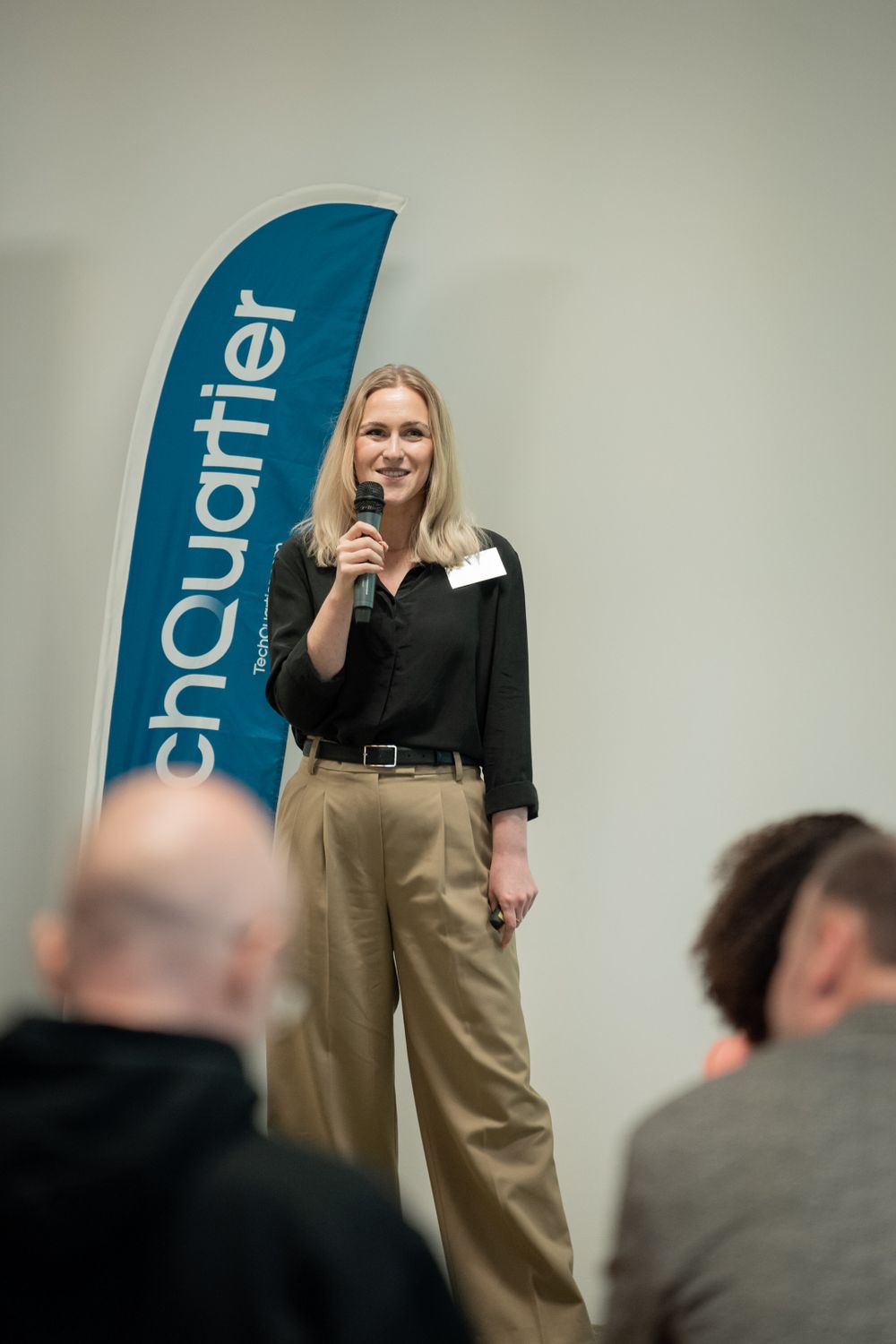 A presenter speaking at a corporate event with a blue TechQuartier banner in the background.