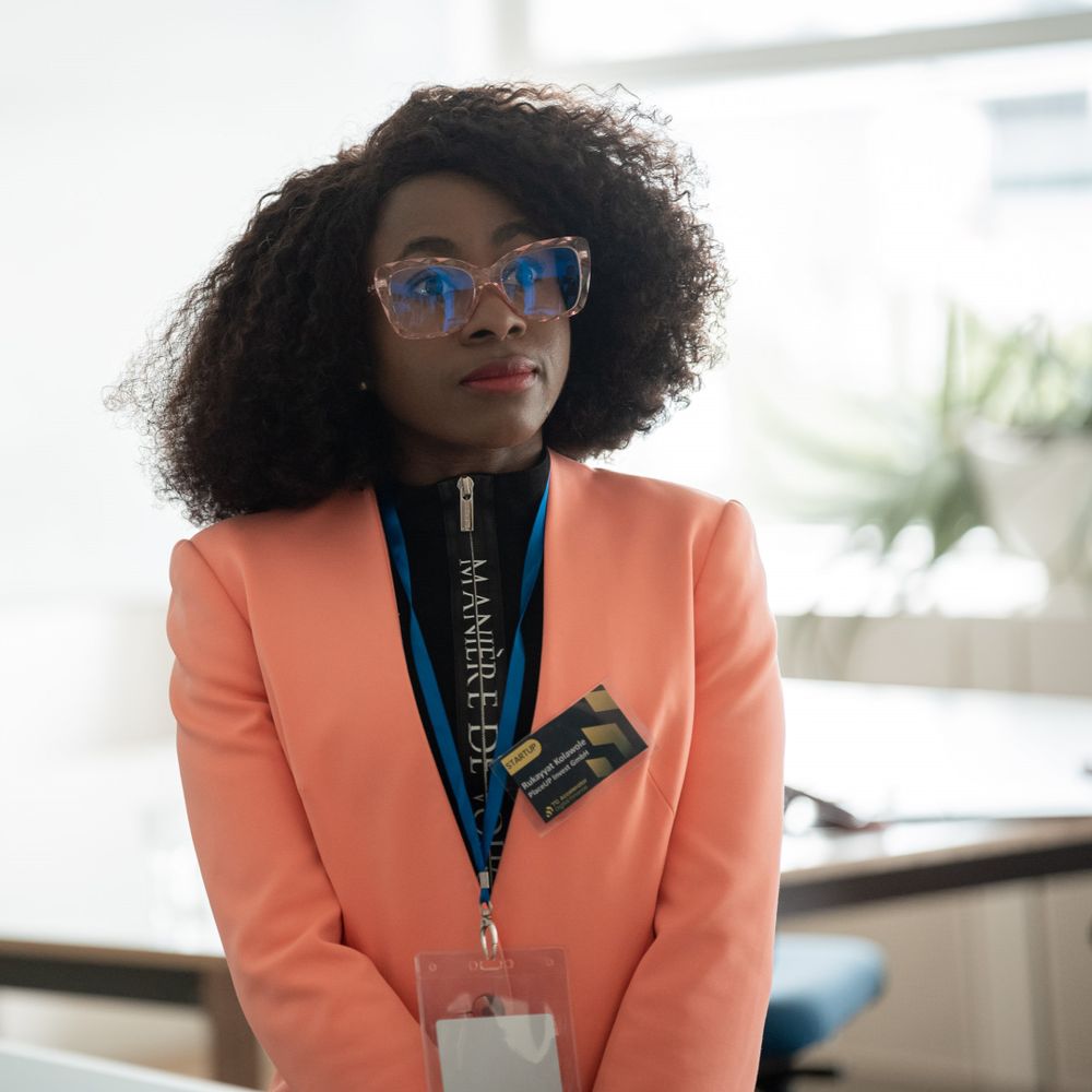 Professional wearing an orange blazer, blue glasses and name tag lanyard in an office setting.