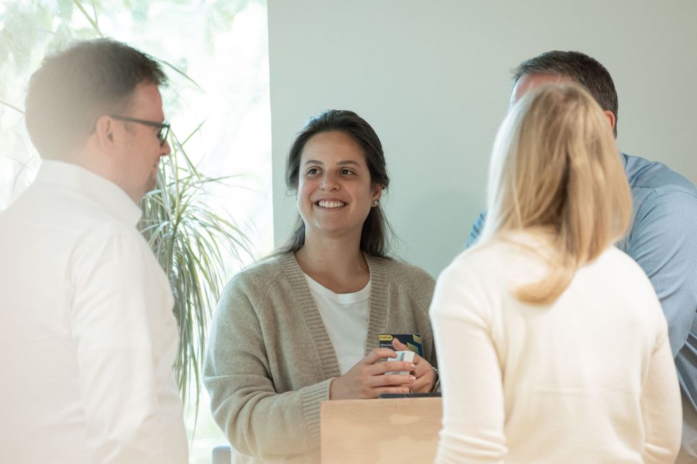 A small group engaged in casual conversation in a bright, plant-decorated office space.