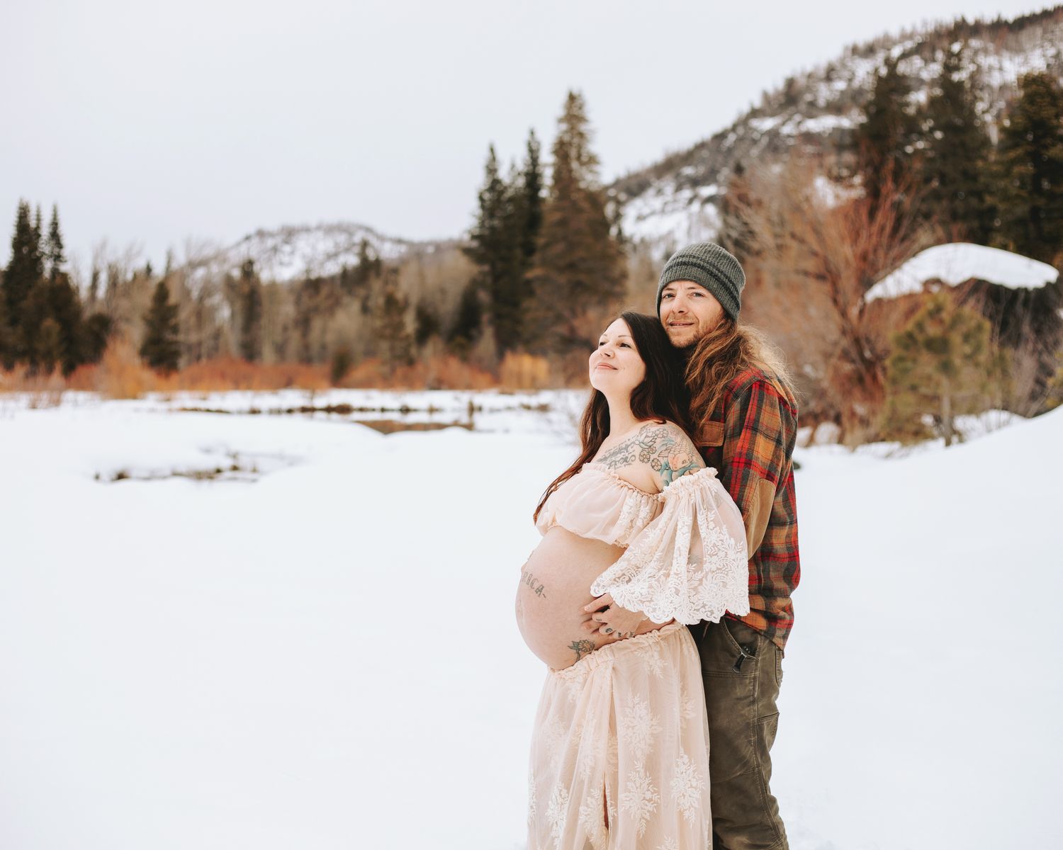 Beautiful tattooed pregnant mom showing off bare baby bump with her partner during Lake Tahoe winter maternity shoot