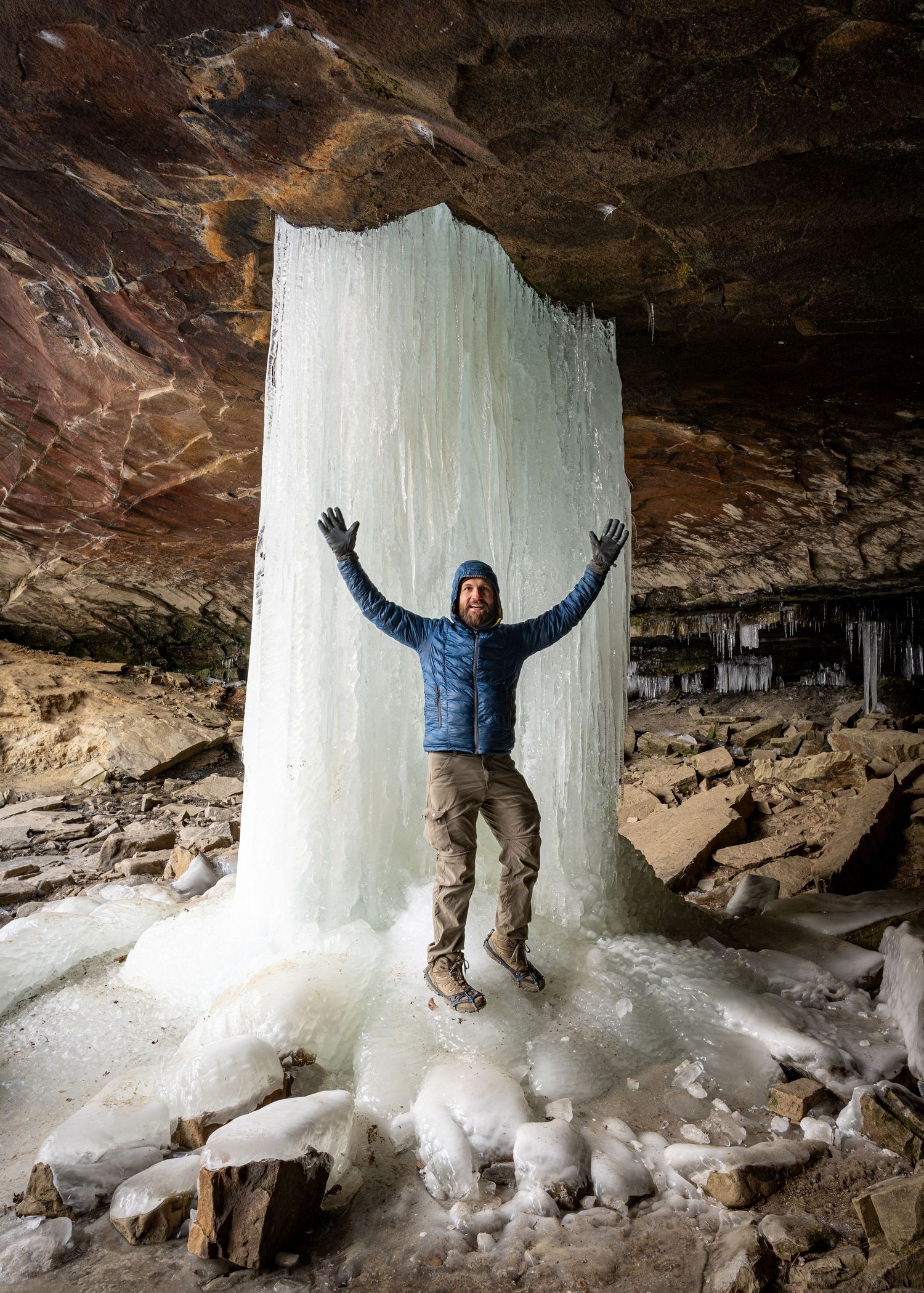 Huge frozen waterfall coming out of a hole in a rock overhang with AR nature photographer, Alex Kent standing in front.