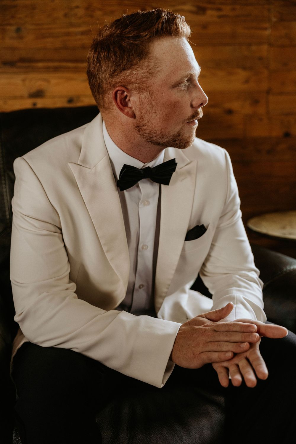 Groom in white tuxedo jacket and black bow tie sits pensively in formal wedding attire.