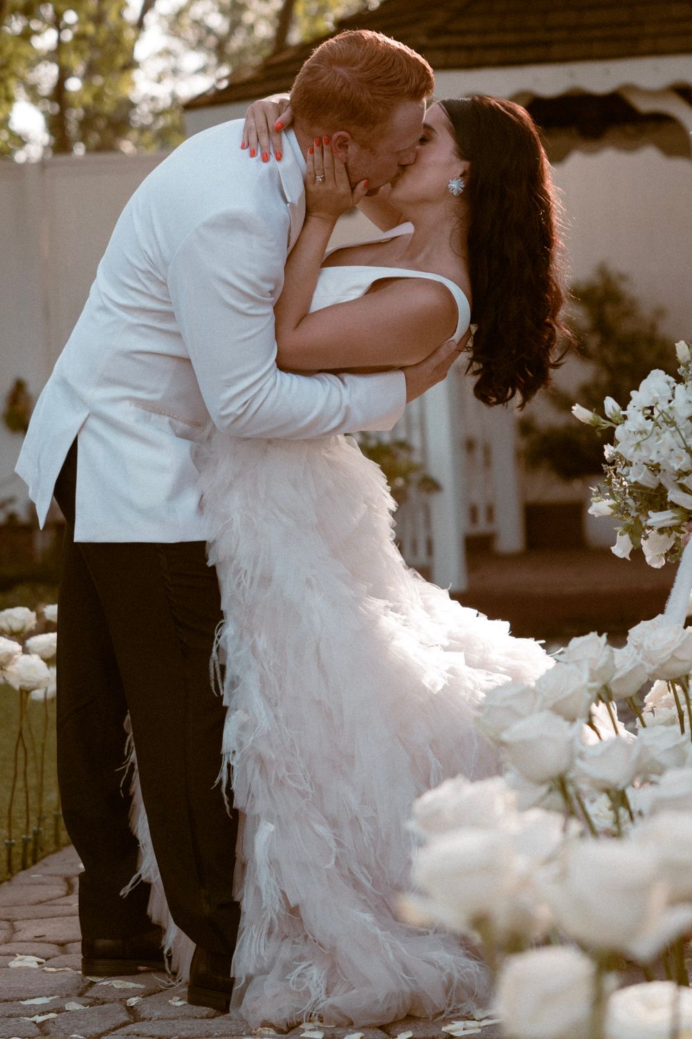 Romantic dip kiss in elegant white wedding attire surrounded by floral decorations.