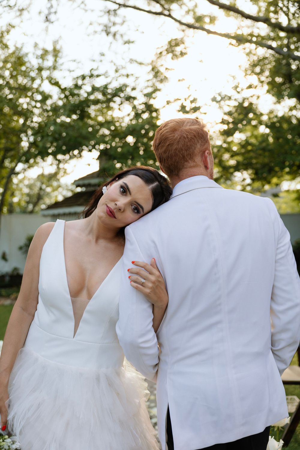 Tender moment between newlyweds in matching white attire against garden backdrop.