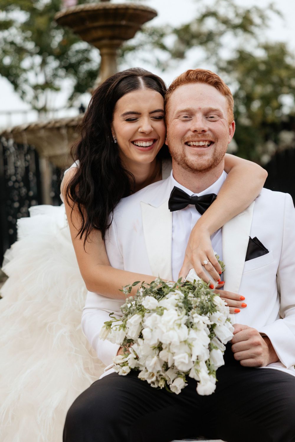 Joyful wedding couple shares intimate moment with white bouquet against fountain backdrop.