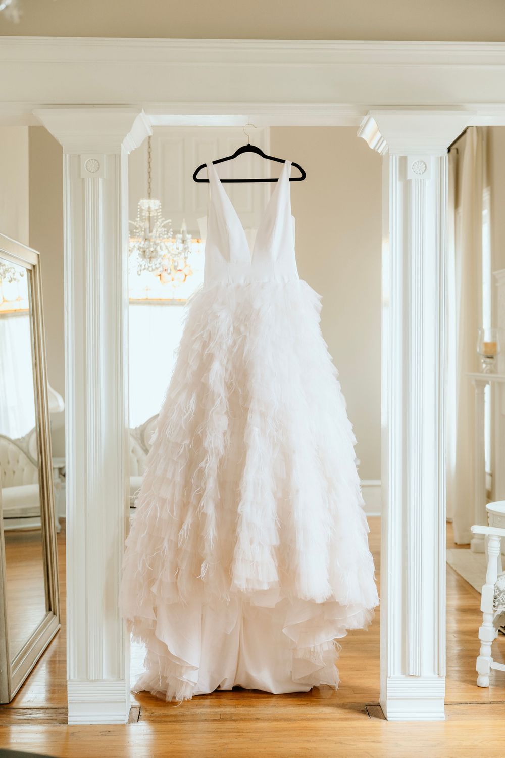 White wedding gown with feathered details hangs elegantly between classic white columns.
