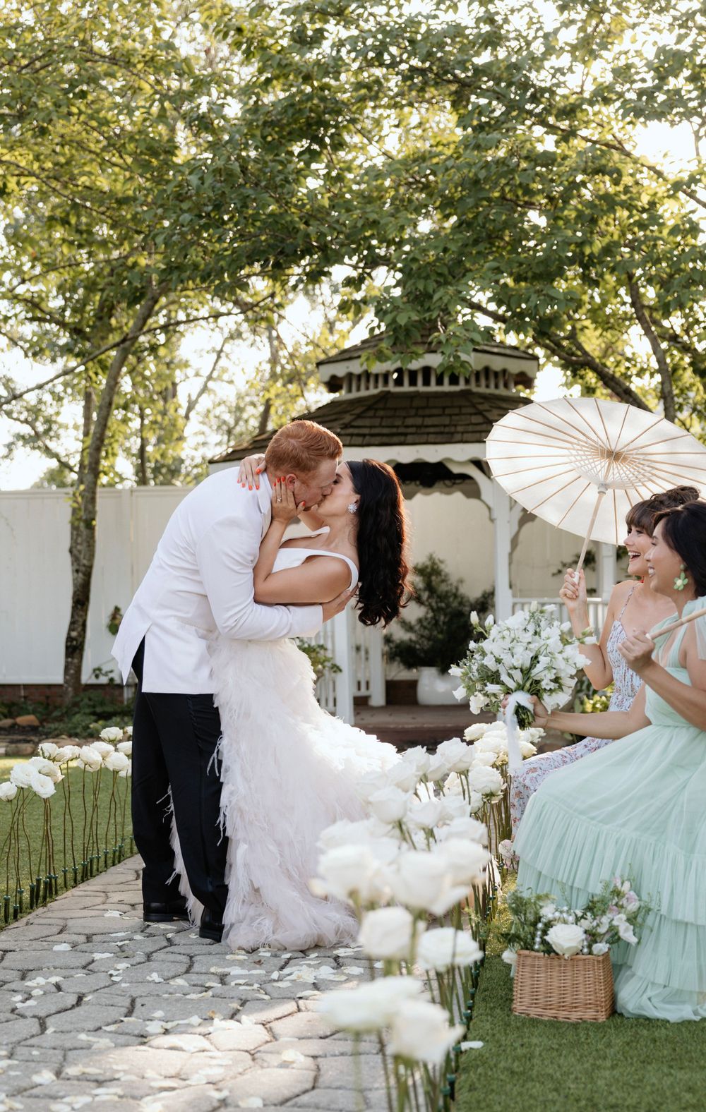 Romantic wedding kiss at an outdoor gazebo ceremony decorated with white flowers and parasols.
