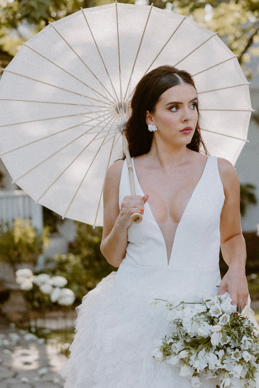 Bride in plunging white gown poses with parasol and bouquet in garden setting.